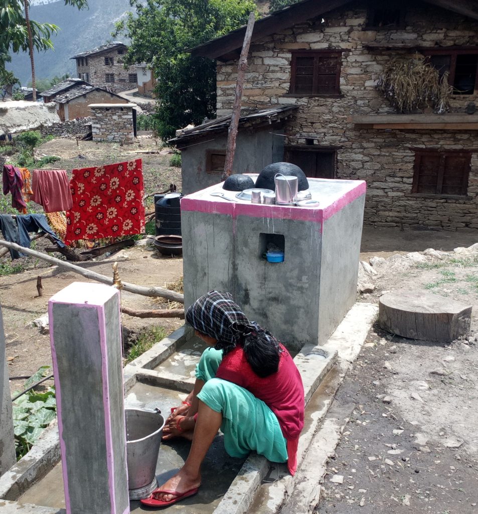 Woman living in rural area of Nepal is washing reusable sanitary pad using private tap in her yard. Houses, trees and mountain are in the background.