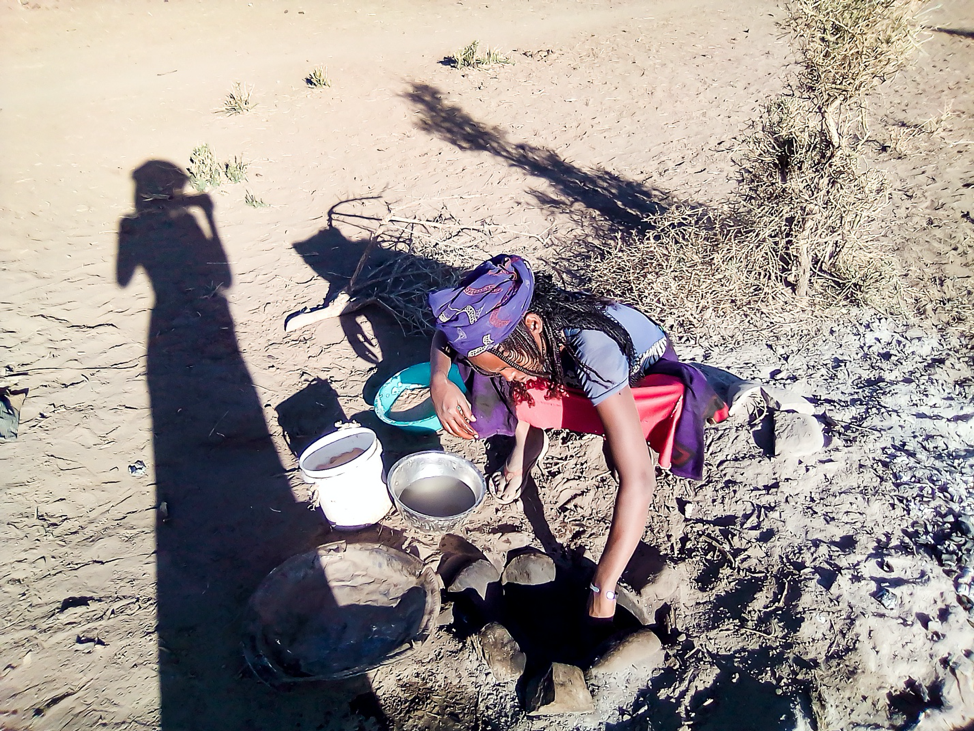 Shadow of person taking the photo on left side. They are photographing of young woman crouched down by a hotstone oven which is dug into the earth. She has various pans and pots around her with ingredients for Mulfie. It is set in the desert in Afar, Ethiopia.