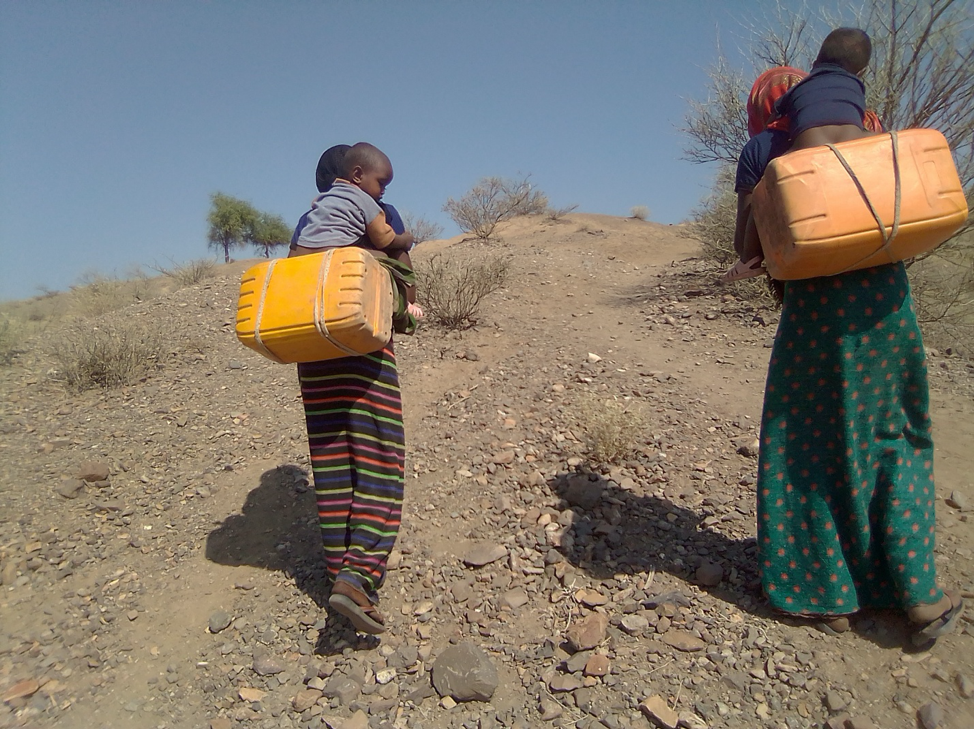 Two women walking away from the camera up a dusty hill with a few dry trees. The women each have a large yellow square water container strapped to theirs backs as well as a small child each sat on top of the can and holding the women around the shoulders..