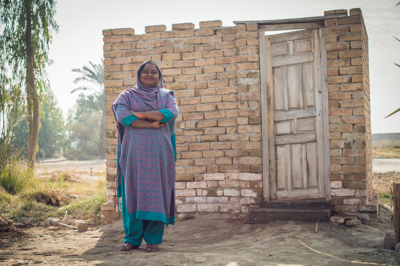 Shazia Qasim, 24, in front of the washroom in her home in the village of Chaoni, District Muzaffargarh, Province Punjab, Pakistan, December 2017. She stands with her arms folded and looking happy.