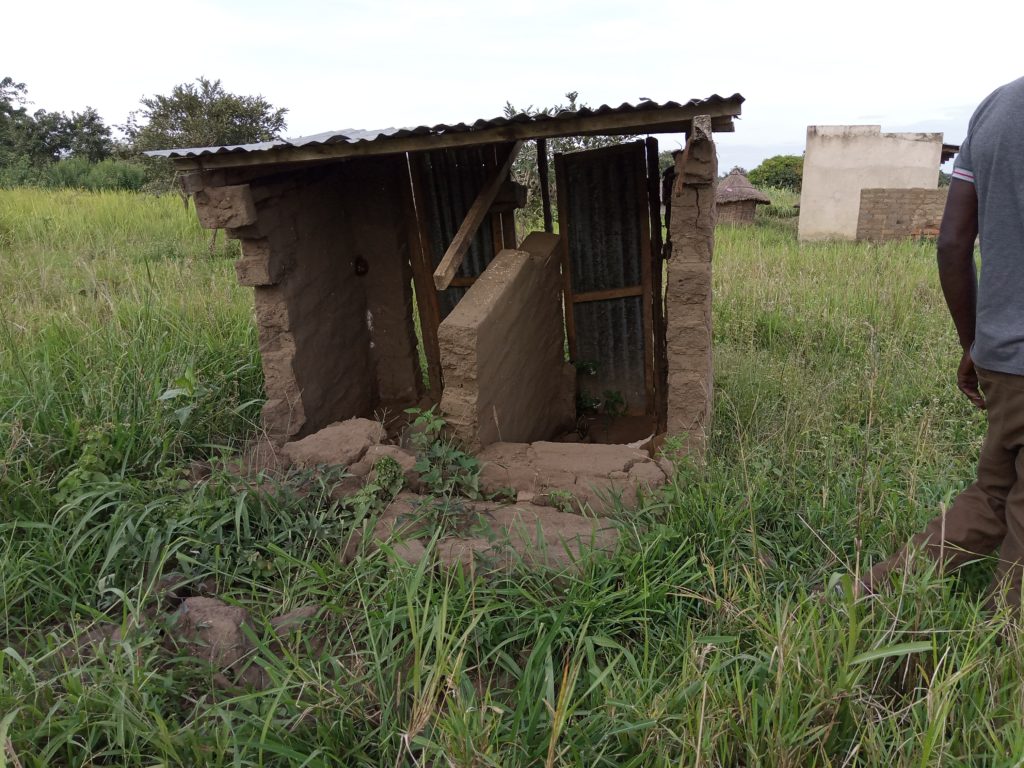 A brick-built latrine with a corrugated iron roof, in a green field. One side has collapsed and the roof is at an angle.