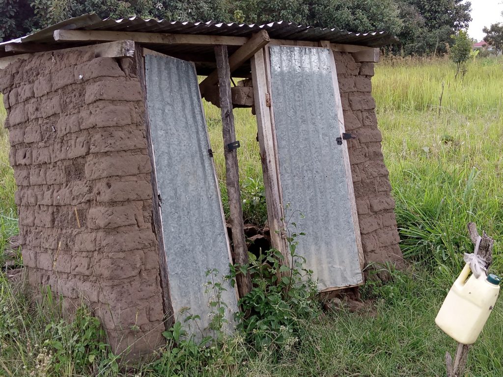A brick-built latrine with corrugated iron roof and doors sinks back into the ground. There is a handwashing station outside.