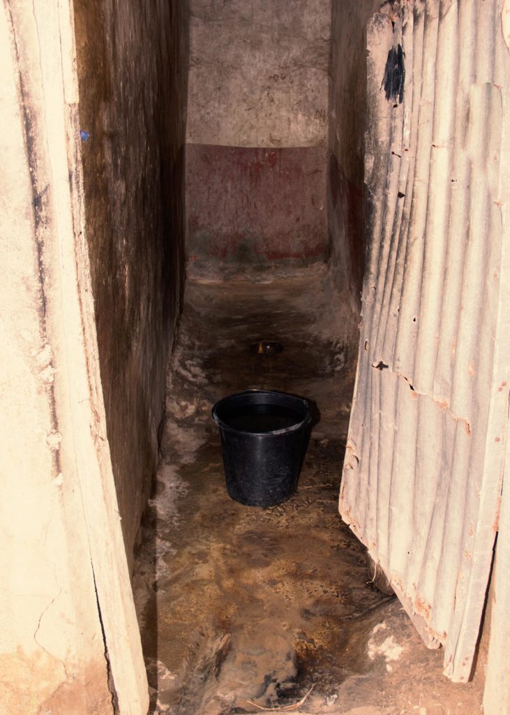 A white corrugated iron door opens to a narrow concrete room which is dusty and dirty. There is a black bucket in front of a small hole in the ground. This serves as a toilet and bathroom.