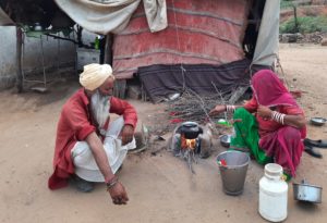 A man and woman from Kalbelia community crouch either side of a fire. The woman is heating a pot.