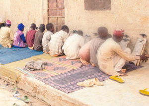 10 Children sit facing a wall reading and writing. On the left side of the photo 3 adults sit watching them.
