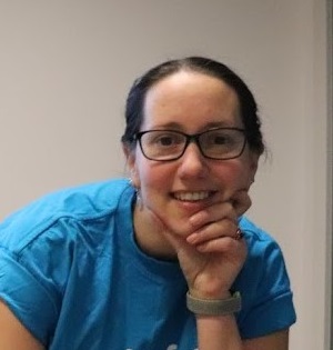 Esther rests her face on one hand and smiles at the camera. She is wearing glasses and a blue t-shirt.