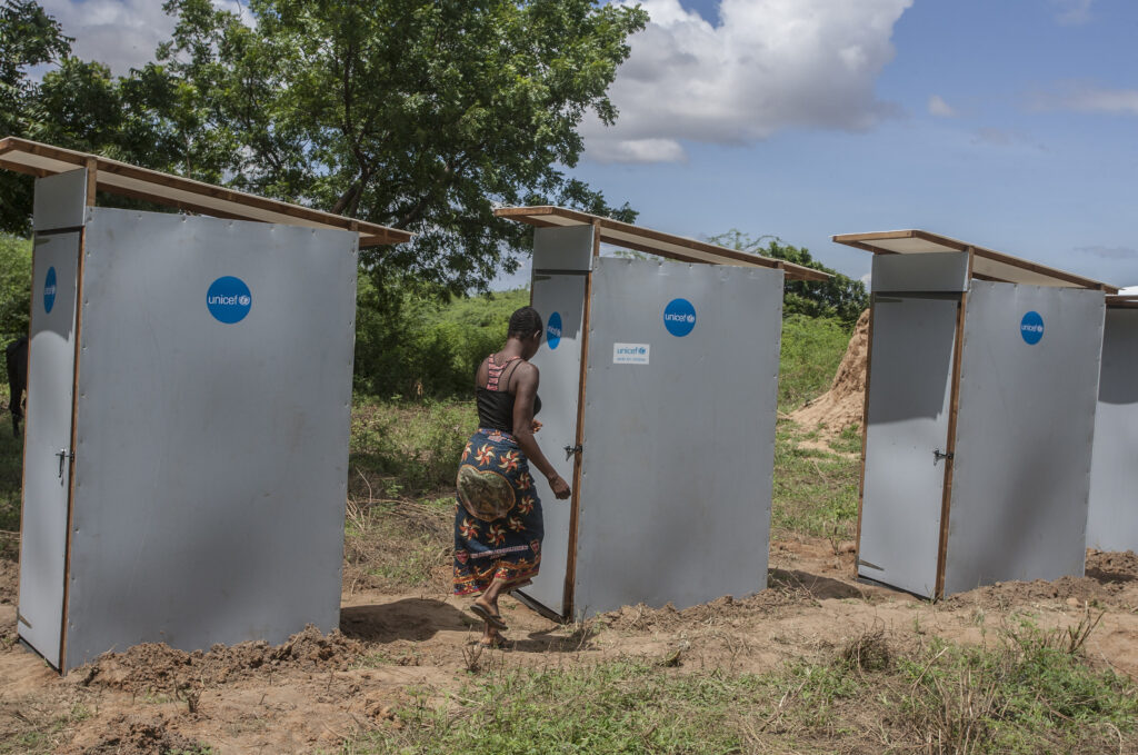 Three toilet cabins made from wood and sheet metal are in the mid-distance. Blue UNICEF stickers are on each cabin. A woman is walking towards the cabin in the centre of the photo, she is about to enter. There is a large tree in the background and area looks dry and rural. 